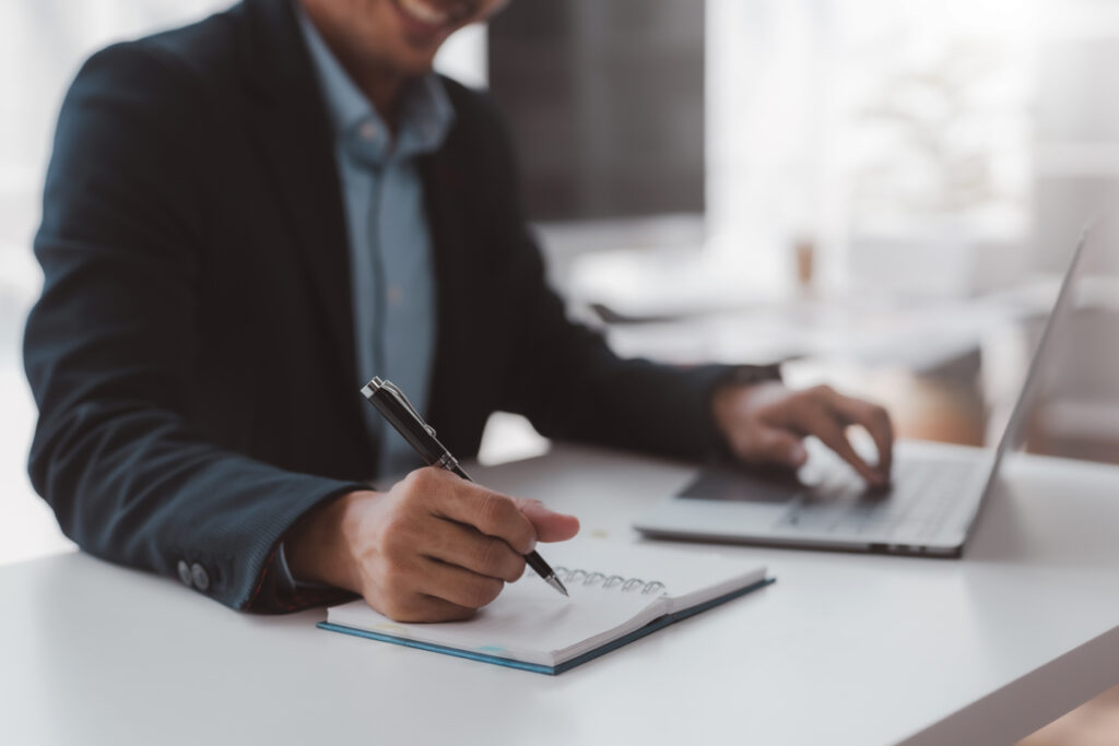 Closeup shot of businessman hand using pen and writing in notebook, presentation, working project at workplace. Man taking notes, typing on keyboard.
