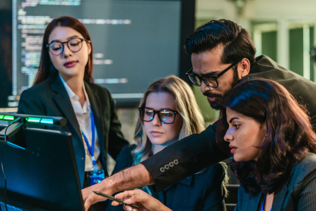 Professionals collaborate in tech environment. Senior members coaching, mentoring, guiding colleagues through data analytics and software development. Knowledge sharing, leadership, technical growth