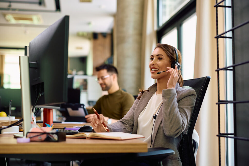 Happy woman talking via headset while having conference call on desktop PC int he office.
