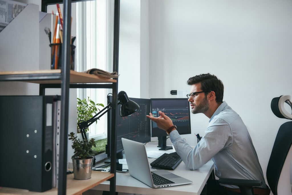 Really Side view of surprised young male stock market broker analyzing a graphs on computer and gesturing while sitting in his modern office. Stock exchange. Trade concept. Investment concept
