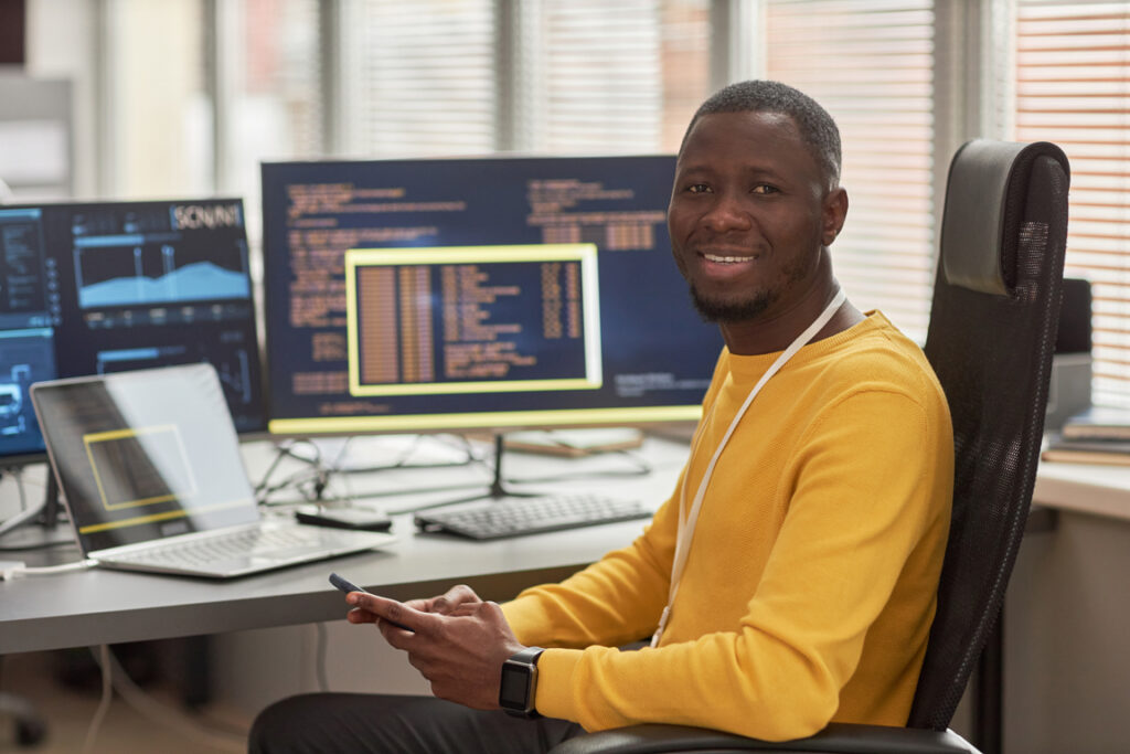 Portrait of smiling black man looking at camera while sitting at workplace in software development company against computer screens with code