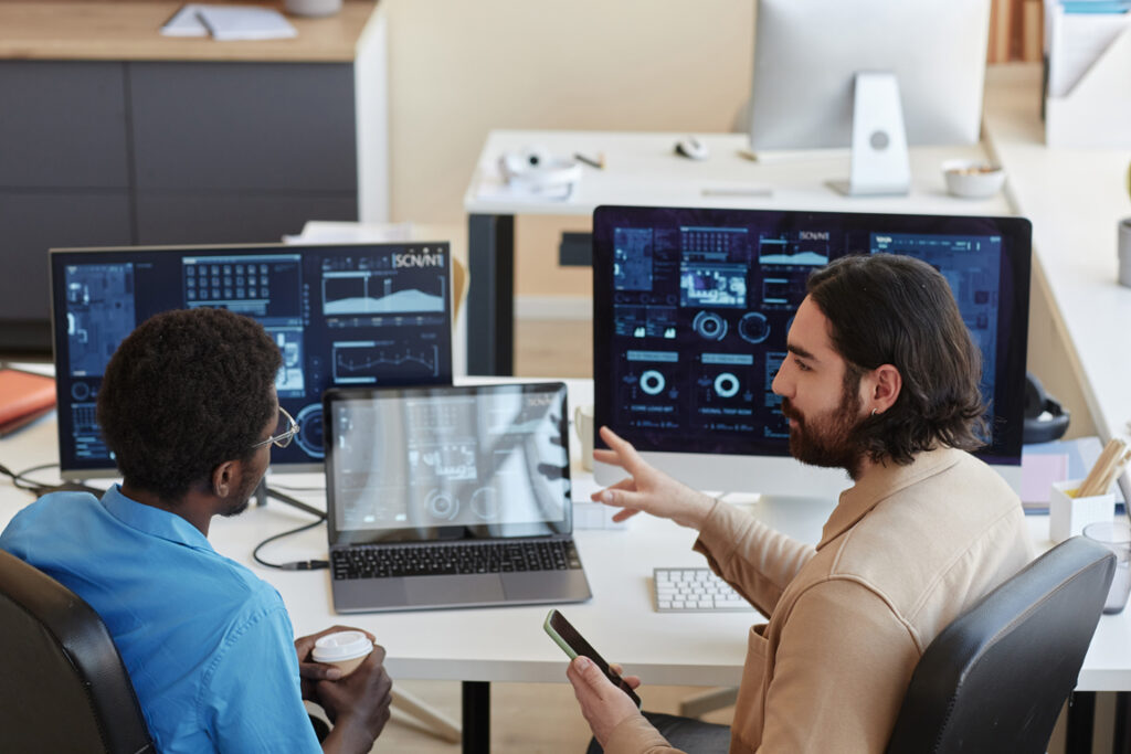 Young confident IT engineer explaining graphic data on computer screens to his colleague while looking at him during working meeting