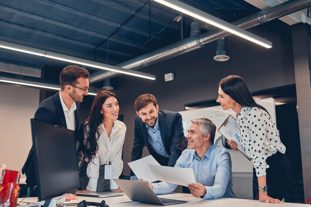 Smiling teammates working in modern office at desk while talking about new project with mature boss